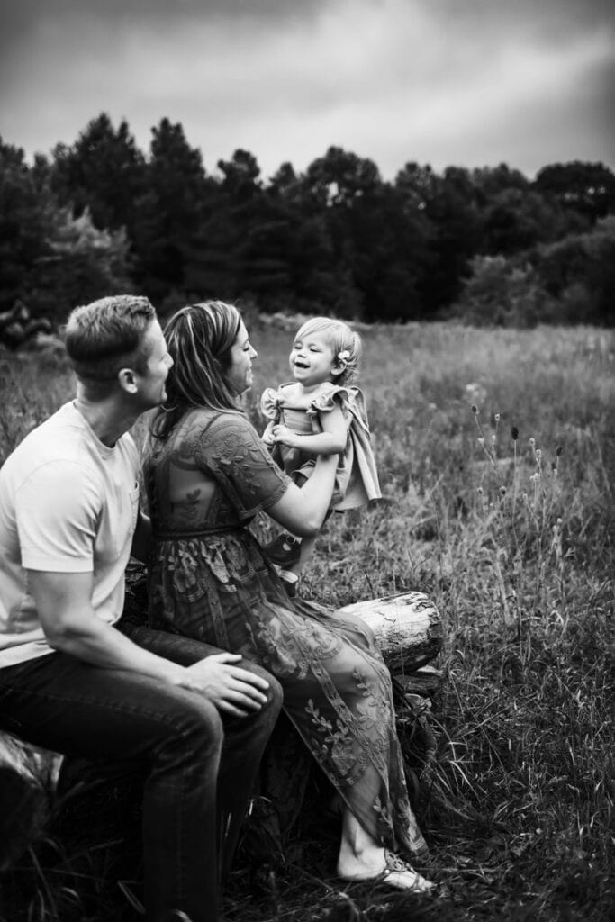 Family Photographer, a young family sits together in a field, mom holds up her baby