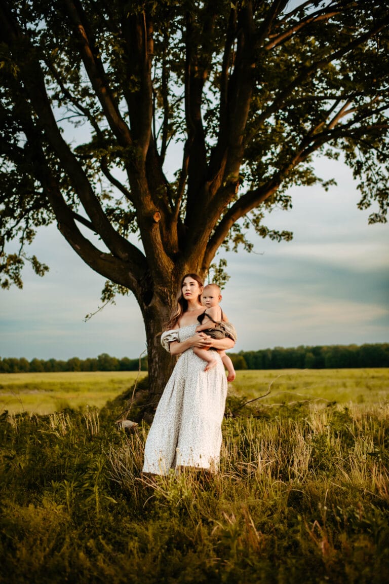 Family Photographer, a woman holds her baby beneath the a large lone tree in a field